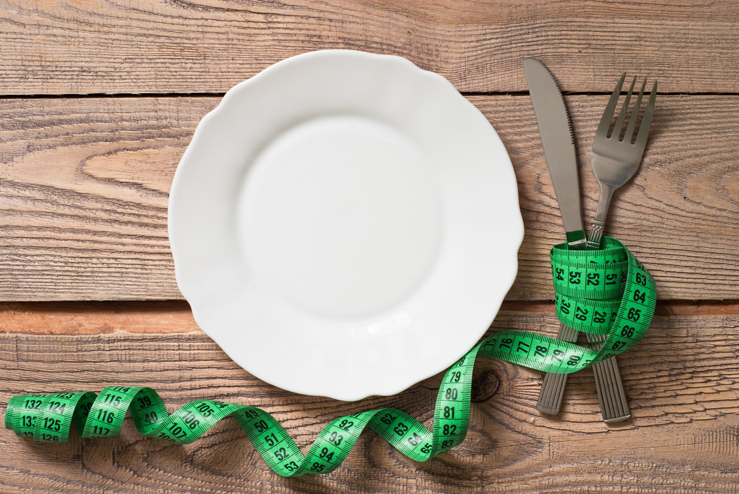 empty white dinner plate on a wood table with a green measurement tape around a knife and a fork